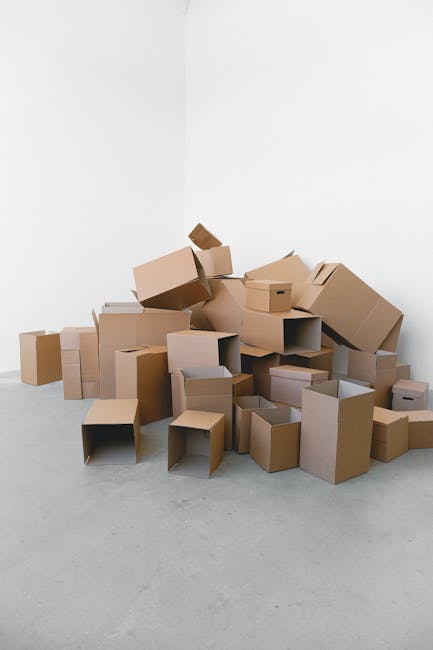 A large pile of cardboard boxes of various sizes, some with open flaps, stacked against a white wall in a room with a light-colored floor. The boxes are in different states of packing, with some appea