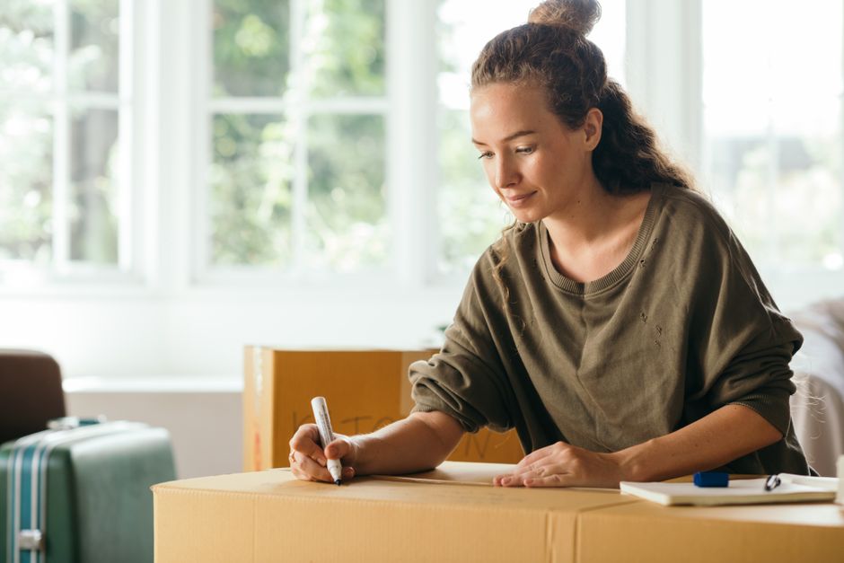 A young woman with curly hair tied in a bun, wearing a dark green long-sleeved shirt, is sitting at a table inside a well-lit room with large windows and a wooden door in the background. She is surrou