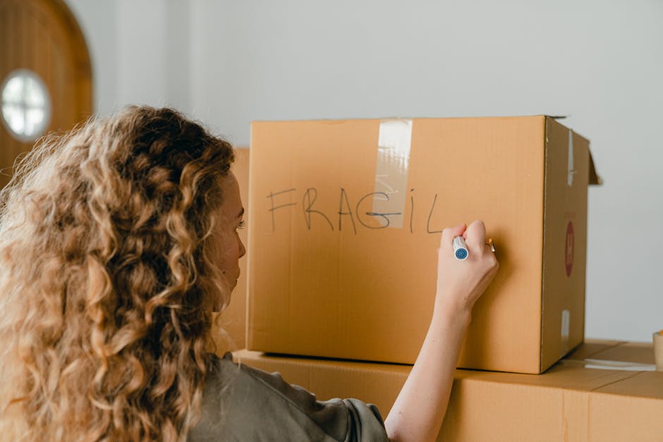 A person wearing a white t-shirt and blue jeans is seen packing a fragile household item, specifically a glass bowl, into a corrugated cardboard box lined with protective materials. Their hands are ca