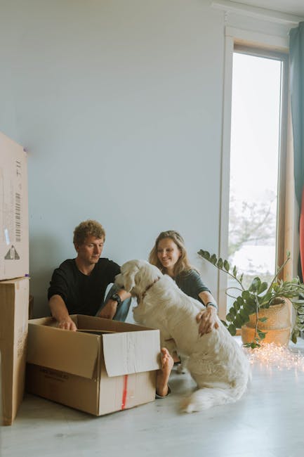 A family preparing to move into a new home, with a woman opening a cardboard box containing a small puppy inside, while a man and a young girl observe and assist. The scene is set in a living room wit