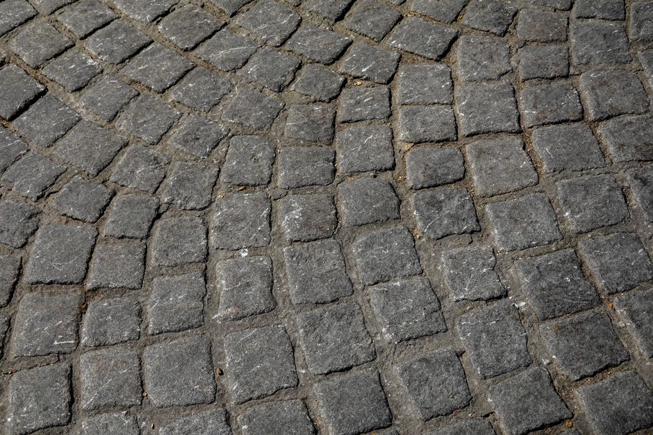 Close-up view of a cobblestone pavement with irregularly shaped, dark grey stones arranged in a circular pattern, showing signs of wear and slight discoloration. The surface appears textured with visi