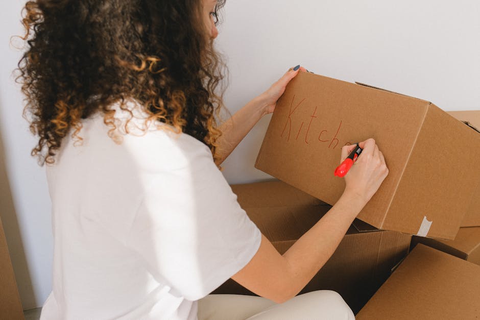 A cardboard box labeled 'Clothes' placed on a windowsill inside a room, with dark curtains partially open revealing a window that shows an exterior brick wall. Next to the box, there is a roll of whit