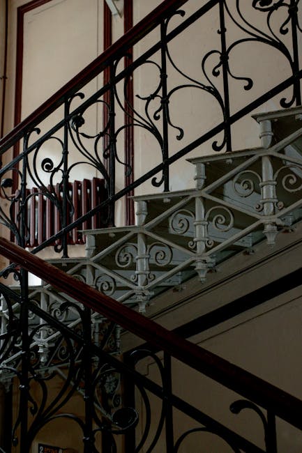 A close-up view of a staircase inside a residential property, featuring a wooden handrail and black metal balusters. The staircase has a strip of red and black patterned carpet runners covering the st